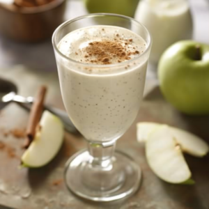 A creamy Cherry Cheer smoothie bursting with goodness, elegantly topped with a sprinkle of cinnamon. Surrounding it, fresh sliced green apples, a whole crisp green apple, and aromatic cinnamon sticks rest on a rustic wooden surface. A cheerful cherry garnish adds a vibrant pop of color to this refreshing and health-boosting treat. In the backdrop, another glass of our energizing Cherry Cheer awaits—just one of the many eco-friendly delights from Zestful Blends!