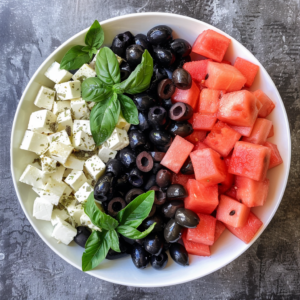 Alt text: Dive into our Watermelon & Feta Salad, a vibrant medley bursting with goodness! This radiant bowl features perfectly cubed feta cheese, succulent black olives, and juicy watermelon, arranged in eye-catching vertical sections. Fresh basil leaves crown the salad, adding a touch of aromatic elegance. Presented against a textured dark backdrop, this delight showcases the freshness and health benefits that define Zestful Blends. Savor every bite while supporting eco-friendly choices!