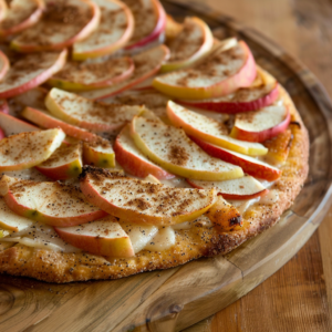 A tantalizing close-up of our Apple Cinnamon Delicacy Pizza, bursting with goodness! This vibrant creation features thinly sliced apples and a sprinkle of cinnamon sugar atop a golden brown crust. Presented on an eco-friendly wooden serving board, this delightful dish embodies the freshness and health benefits Zestful Blends is renowned for. The blurred background ensures all eyes are on this mouthwatering masterpiece. Indulge in a dessert that’s both delicious and wholesome!