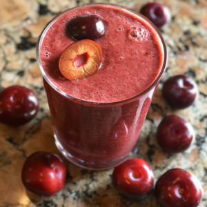 A close-up of our vibrant Cherry Jubilee Smoothie in a sleek glass, garnished with a halved and a whole cherry, exuding freshness. Several plump cherries are playfully scattered around the glass on a speckled countertop. Bursting with goodness, this delightful treat is packed with antioxidants and natural sweetness, ensuring every sip is as healthful as it is delicious. At Zestful Blends, we believe in harnessing the purest ingredients for a tantalizing taste that's kind to both you and the planet.