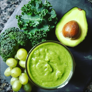 A vibrant Green Grape Oasis Smoothie sits in a glass bowl on a sleek slate surface, exuding healthful radiance and eco-friendly charm. Surrounding the smoothie are fresh ingredients that reflect its bursting vitality: an open avocado with its pit, a bunch of juicy green grapes, crisp kale leaves, and nutrient-packed broccoli florets. Experience the zestful blend of nature's finest with every sip—pure freshness and wellness in one delightful treat.